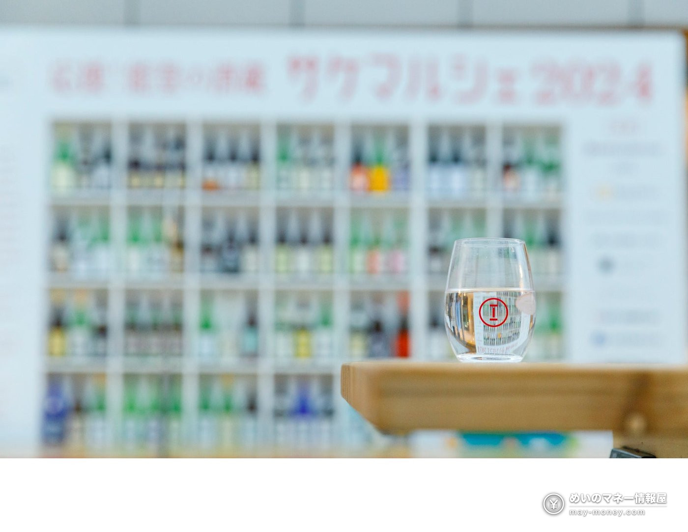 A Sake Marche logo glass on a counter with shelves of sake bottles behind it