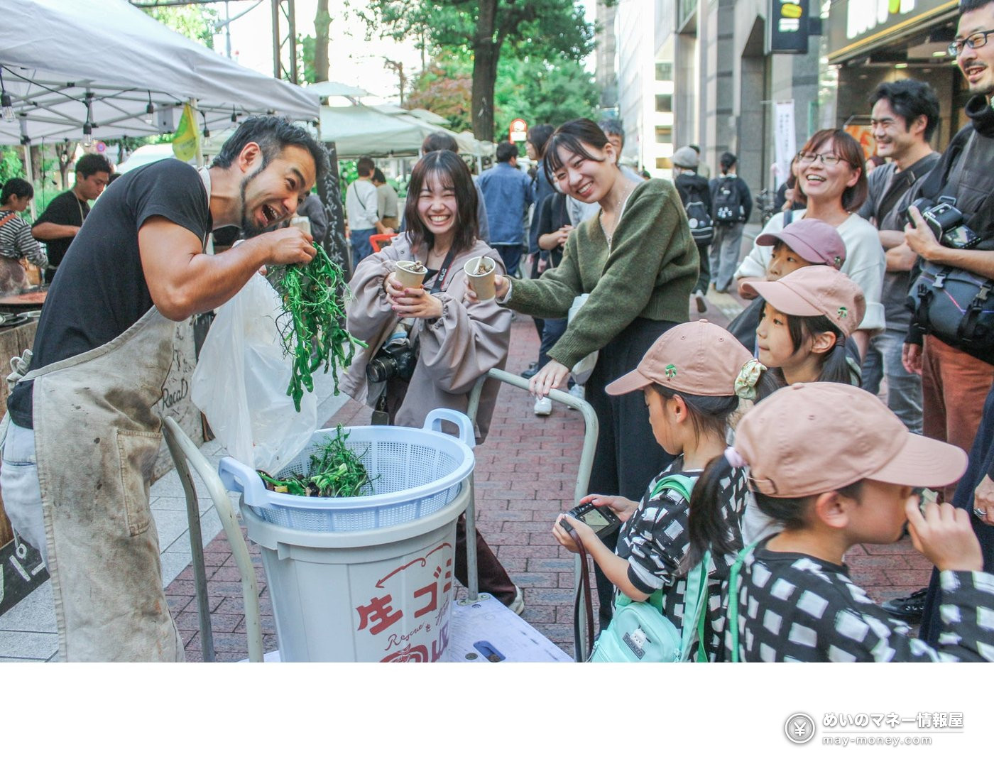 男性把厨余倒进写着生ゴミ的桶里，周围的孩子和大人在笑