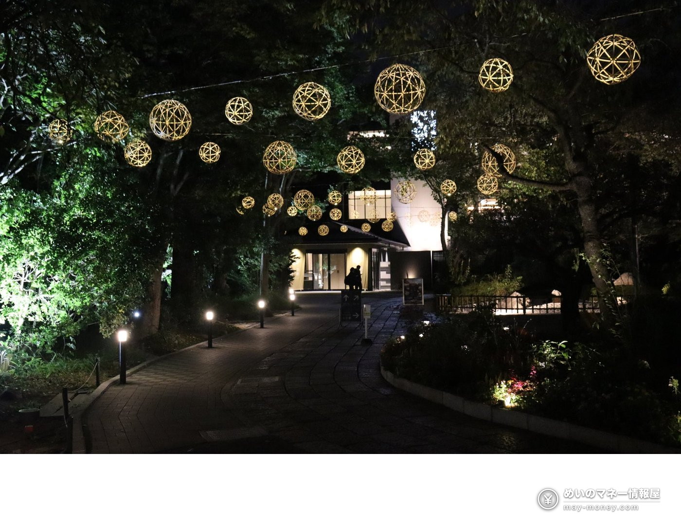Bamboo ball lanterns hanging from trees over a path at night, with a bright building behind