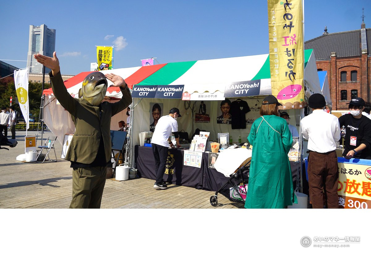 魚の頭のかぶり物をした人物が両腕を広げ、背後に出展テントが並ぶ会場の様子