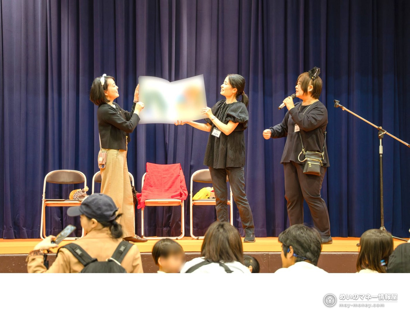 Three women reading a large picture book aloud on stage to an audience