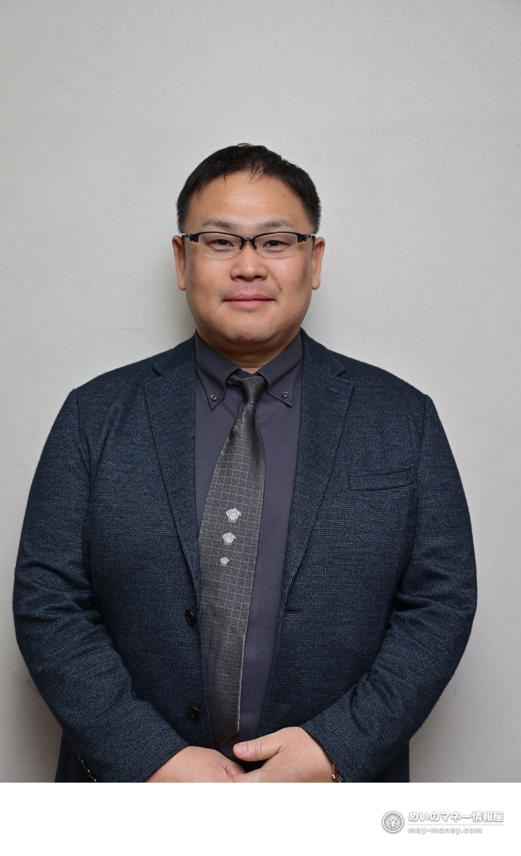 A man in glasses and a suit standing in front of a grey wall