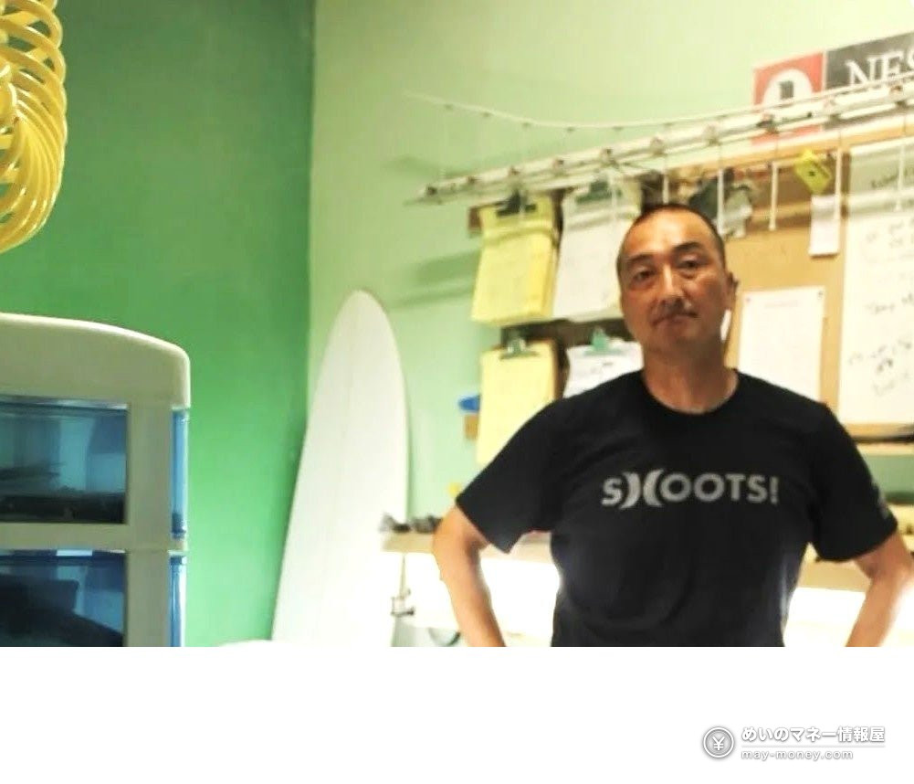 サーフボードを背に、緑の壁の部屋で腰に手を当てて立つ黒いTシャツの男性