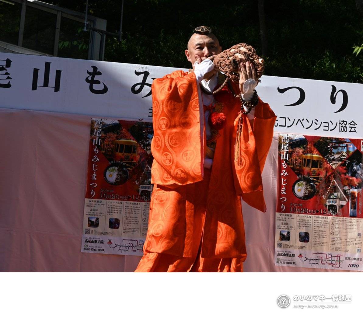 身穿橙色装束的山伏在写有高尾山もみじまつり的幕布前吹法螺贝