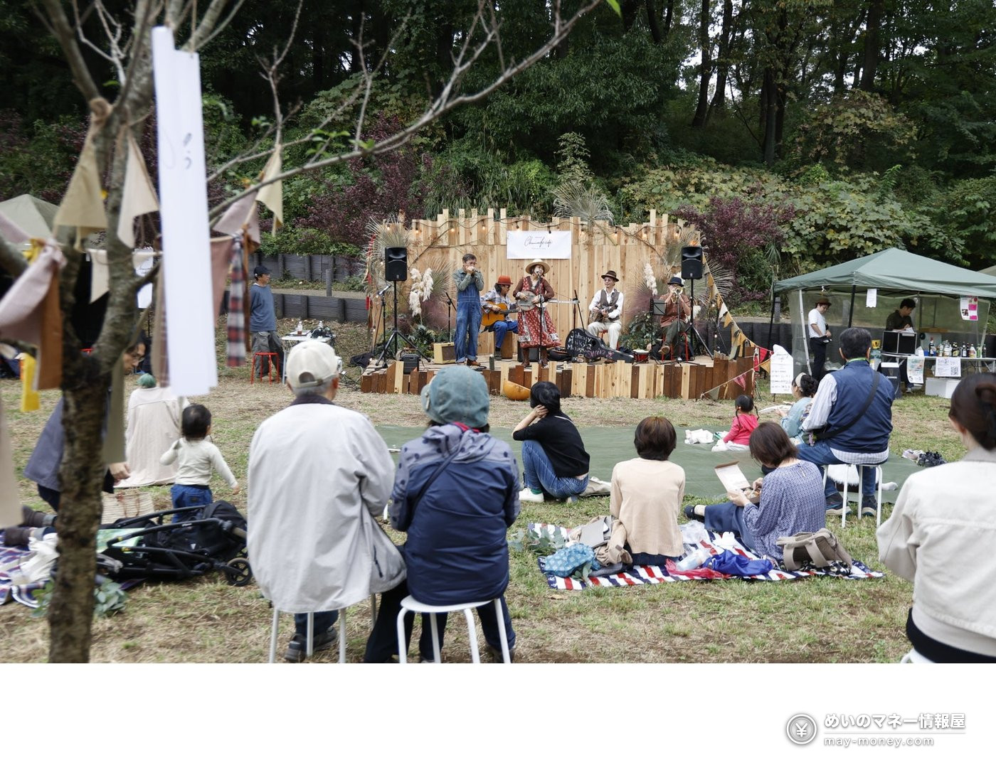 屋外の木製ステージで演奏するバンドと、芝生で聴く観客