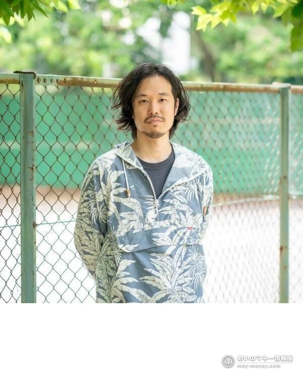 A long-haired man in a floral jacket standing in front of a wire fence