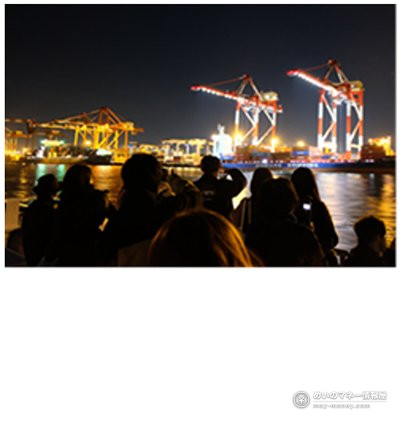 夜の港でライトに照らされたコンテナクレーンを船上から眺める人たち